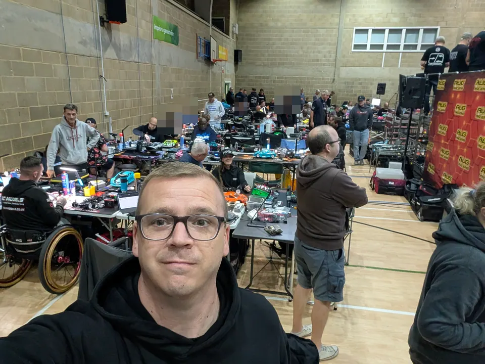 A selfie in a busy sports hall: rows of pit tables behind, racers working on their cars, and the red RugRacers banner on the right.