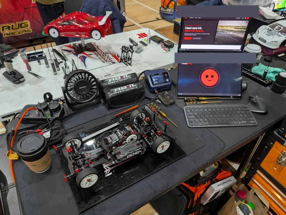 The Xpress MF1 on a black pit mat beside a coffee, a fan and a charger, with a two-screen laptop showing the Marbl Racing home page and the Marbl Pit Crew face.